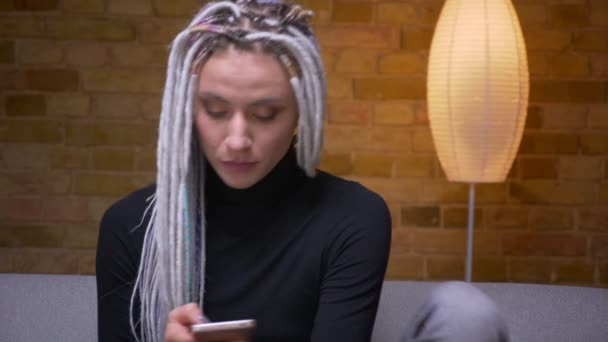 Close-up tiro de jovem atraente caucasiano hipster fêmea com dreadlocks loira mensagens no telefone sentado no sofá dentro de casa em um aconchegante apartamento — Vídeo de Stock