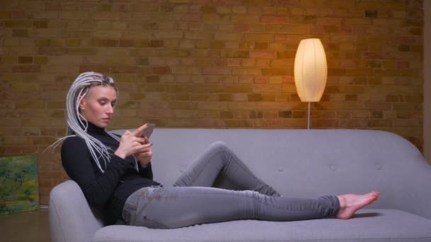居心地の良いアパートのソファに座って携帯電話を使用して若い魅力的な白人ヒップスターの女性のクローズアップサイドビュー撮影 — ストック動画
