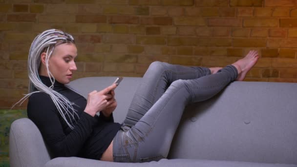 居心地の良いアパートのソファの上に彼女の足を置く携帯電話を使用して若い魅力的な白人ヒップスターの女性のクローズアップサイドビュー撮影 — ストック動画