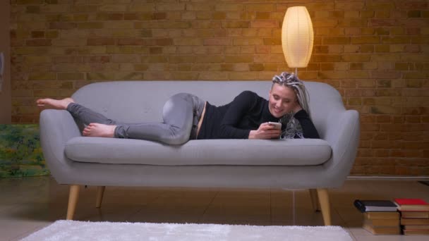 Съемка с близкого расстояния молодой привлекательной кавказки с белокурыми дредами, смотрящими по телефону, опирающимися на руку, лежащую на диване — стоковое видео