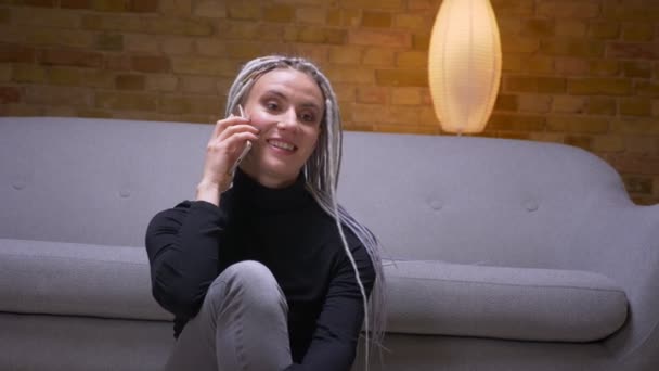 Primo piano sparare di giovane attraente femmina caucasica con dreadlocks bionda chiamando al telefono sorridente felicemente seduto sul pavimento al chiuso — Video Stock