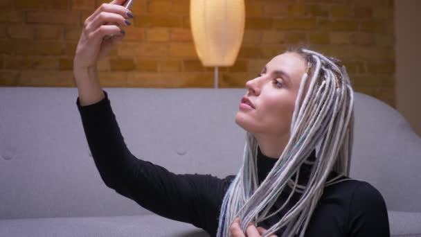 Primer plano de brote de joven atractiva mujer caucásica con rastas rubias tomando selfies en el teléfono sentado en el suelo en interiores — Vídeo de stock