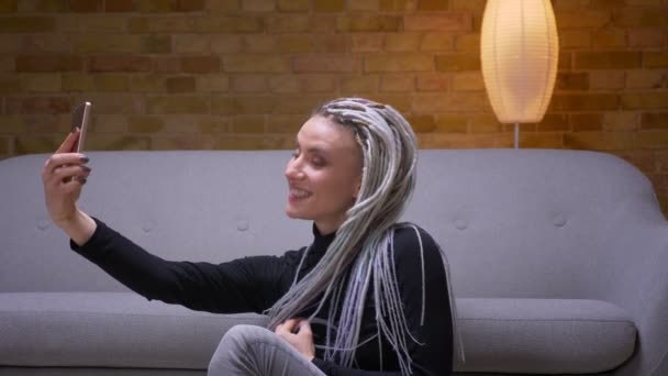 Zbliżenie strzelać z młody atrakcyjny kaukaski żeński z Blondynka dredy biorąc selfie na telefon stwarzające przed kamerą siedzi na podłodze w pomieszczeniu — Wideo stockowe