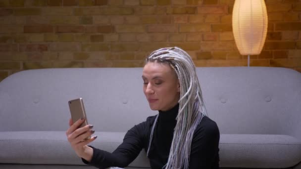 Close-up shoot van jonge aantrekkelijke Kaukasische vrouw met blonde dreadlocks met een video-oproep op de telefoon zittend op de vloer binnenshuis — Stockvideo