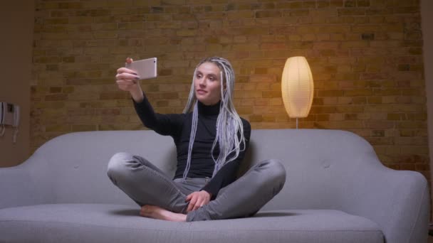 Primer plano de la joven atractiva mujer caucásica con rastas rubias teniendo una videollamada en el teléfono sentado en el sofá en el interior — Vídeos de Stock