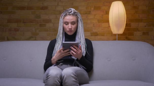 Gros plan pousse de jeune jolie caucasienne hipster femelle à l'aide de la tablette et rire heureux assis sur le canapé à l'intérieur dans un appartement confortable — Video