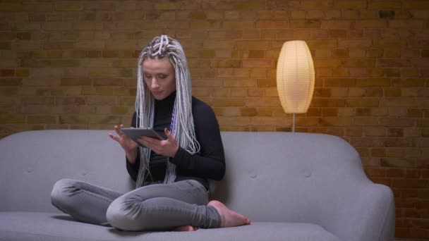 Gros plan pousse de jeune hipster caucasien attrayant femelle naviguant sur la tablette assise sur le canapé à l'intérieur dans un appartement confortable — Video