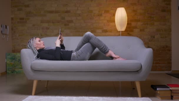 Primer plano de brote de joven atractiva hembra hipster caucásico utilizando la tableta que se encuentra en el sofá en un acogedor apartamento — Vídeos de Stock