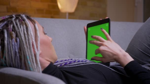 Närbild tillbaka Visa shoot av unga kaukasiska hipster hona med dreadlocks använda tabletten med grön skärm som ligger på soffan inomhus i en mysig lägenhet — Stockvideo