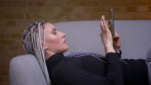 Gros plan pousse de jeune jolie caucasienne hipster femelle à l'aide de la tablette se tournant vers la caméra et souriant joyeusement couché sur le canapé à l'intérieur dans un appartement confortable — Video