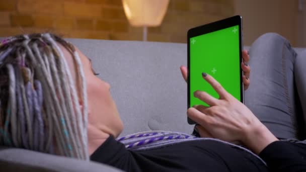 Primer plano de la vista trasera de la sesión de jóvenes caucásicos hipster hembra con rastas utilizando la tableta con pantalla verde con el anuncio en él acostado en el sofá interior en un acogedor apartamento — Vídeos de Stock