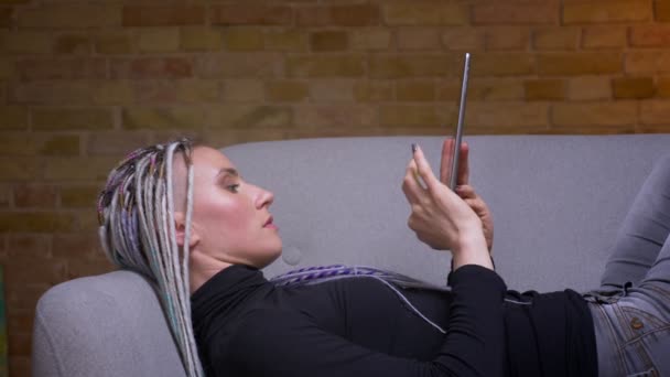 Closeup shoot of young attractive caucasian hipster female with dreadlocks using the tablet turning to camera and happily cheerfully lying on the couch indoors in a cozy apartment — Stock Video