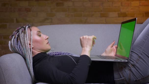 Gros plan pousse de jeune jolie femme hipster caucasienne avec dreadlocks tapant sur l'ordinateur portable avec écran chromatique vert couché sur le canapé à l'intérieur dans un appartement confortable — Video