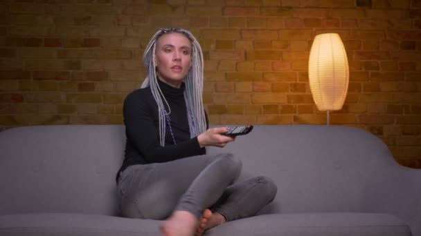 Närbild shoot av unga attraktiva kaukasiska hipster kvinnlig tittar på TV och använder fjärrkontroll sitter på soffan inomhus i en mysig lägenhet — Stockvideo