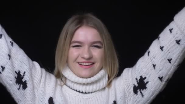 Съемки крупным планом молодой белой девушки в свитере, взволнованной и улыбающейся, счастливо смотрящей в камеру на фоне, изолированном на черном — стоковое видео