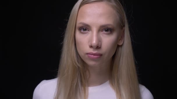 Портрет крупным планом молодой симпатичной блондинки, смотрящей в камеру на фоне, изолированном на черном — стоковое видео