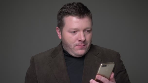 Brunette middle-aged businessman in suit working with phone gets extremely amused and contented on gray background. — Stock Video