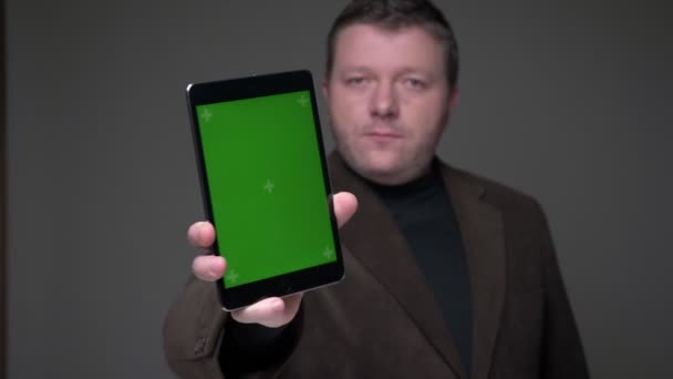 スーツを着たブルネット中年のビジネスマンは、灰色の背景に思慮深くタブレットの緑色の画面を示しています. — ストック動画