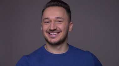 Genç yakışıklı beyaz adamın heyecanla gülümseyen yakın çekimi, gri zemini izole edilmiş arka planlı kameraya bakarak