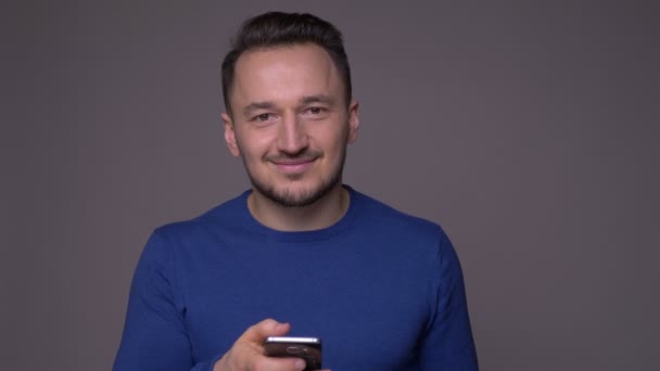 Zbliżenie strzelać młodego przystojnego kaukaski mężczyzna za pomocą telefonu i uśmiechnięty patrząc na kamerę z tła izolowane na szarym — Wideo stockowe