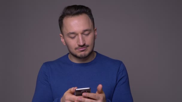 Съемки крупным планом молодого красивого кавказца, обменивающегося сообщениями по телефону с изолированным на сером фоне — стоковое видео