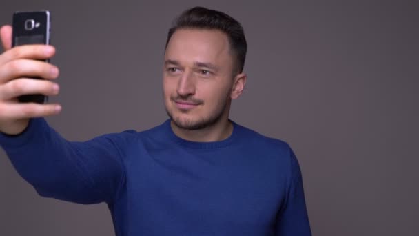 Zbliżenie strzelać młodego przystojnego kaukaski mężczyzna biorąc selfie na telefon i uśmiechnięty z tłem na białym tle na szarym — Wideo stockowe