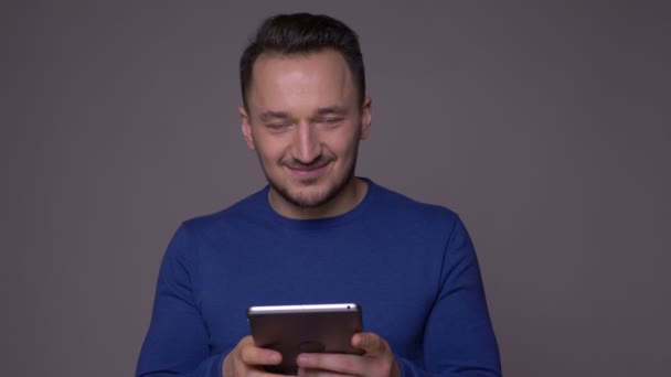 Primer plano de brote de joven guapo hombre caucásico utilizando la tableta sonriendo y mirando a la cámara con fondo aislado en gris — Vídeos de Stock