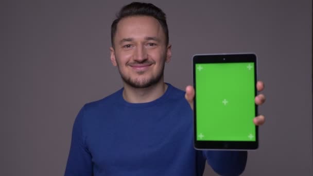 Disparo de primer plano de hombre caucásico guapo joven usando la tableta y mostrando la pantalla de clave de croma verde a la cámara con fondo aislado en gris — Vídeos de Stock