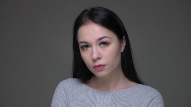 Съемки крупным планом молодой грустной брюнетки кавказской женщины, расстроенной и подавленной, смотрящей прямо в камеру с изолированным на сером фоне — стоковое видео