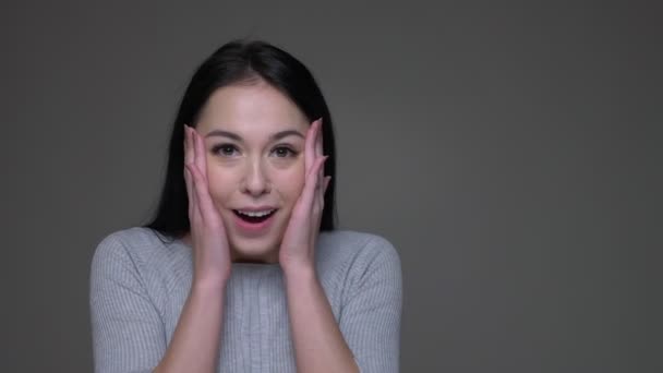 Primer plano de la joven guapa morena caucásica hembra sorprendida y sonriente mientras mira directamente a la cámara con el fondo aislado en gris — Vídeos de Stock