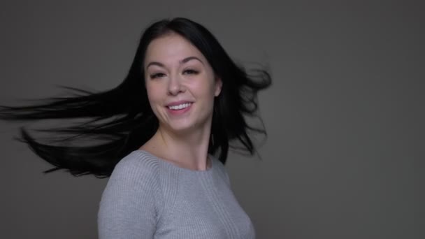 Nahaufnahme Shooting der jungen hübschen brünetten kaukasischen Frau, die sich mit flatternden Haaren umdreht und den Daumen nach oben zeigt, mit isoliertem Hintergrund auf grau — Stockvideo