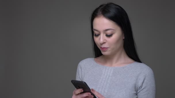 Primer plano de brote de joven guapa morena caucásica femenina utilizando el teléfono con fondo aislado en gris — Vídeo de stock