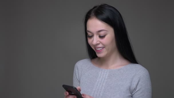 Крупный план съемки молодой симпатичной брюнетки кавказской женщины, просматривающей социальные сети по телефону и реагирующей на фоне изолированного на сером — стоковое видео