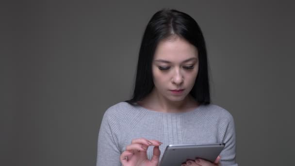 Primo piano di scatto di giovane bella bruna caucasica messaggistica femminile sul tablet guardando la fotocamera con sfondo isolato su grigio — Video Stock