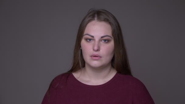 若い悲しい太りすぎの白人女性のクローズアップポーターは、灰色で隔離された背景を持つカメラを見て動揺し、落ち込んでいる — ストック動画