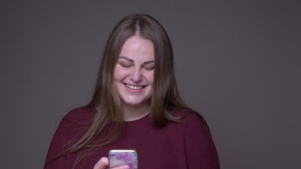 Gros plan portarit de jeune femme caucasienne en surpoids utilisant le téléphone et souriant joyeusement avec fond isolé sur gris — Video