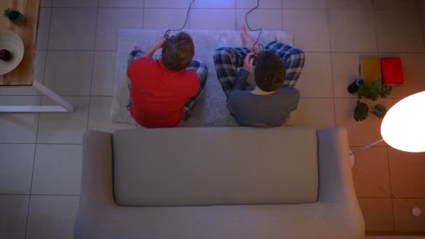 Top shot de dos chicos jóvenes en ropa de dormir jugando videojuego en el suelo y reaccionando emocionalmente en la sala de estar . — Vídeo de stock