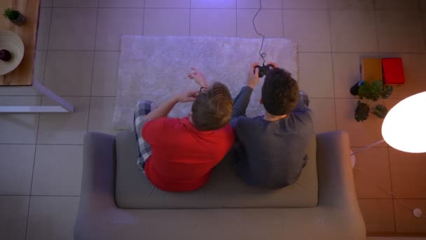 Top shot of two young guys in sleepwear playing videogame and communicating with active gestures in the living room. — Stock Video