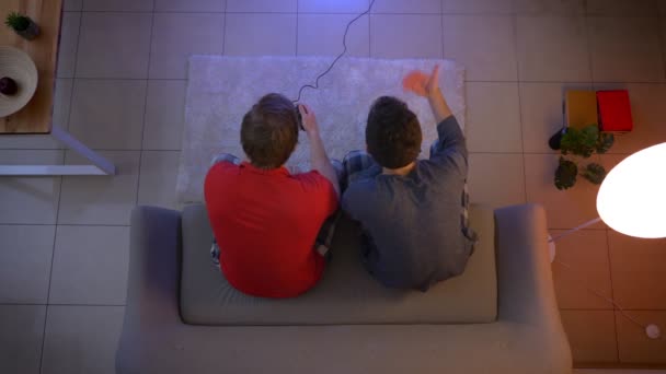 Top shot of guy in sleepwear playing videogame and another one supporting him with active gestures in the living room. — Stock Video