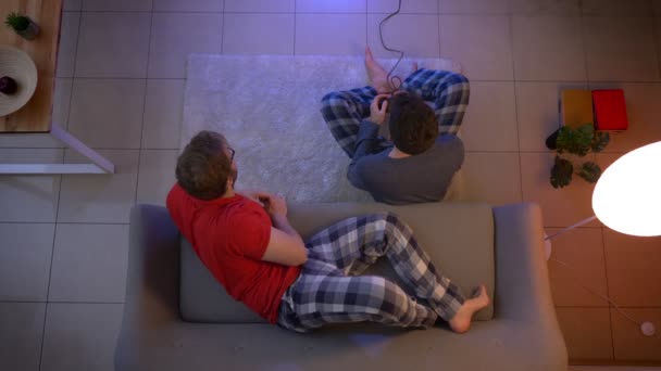 Top shot de chico en ropa de dormir jugando videojuego y otro relajándose en el sofá y hablando con él en la sala de estar . — Vídeos de Stock