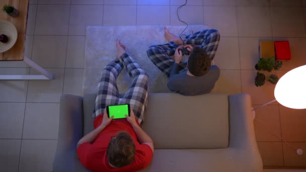Joystick kullanarak video oyunu oynayan ve oturma odasında tablet ile çalışan pijama iki genç çocuklar top çekim. — Stok video