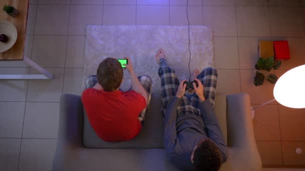 Top shot d'amis en vêtements de nuit jouant au jeu vidéo à l'aide d'un joystick et travaillant avec un smartphone dans le salon . — Video