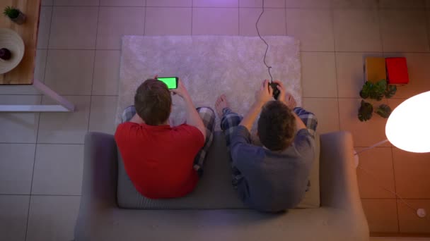Найкращий знімок друзів у сплячому одязі, які грають відеоігри з джойстиком і працюють зі смартфоном у вітальні . — стокове відео