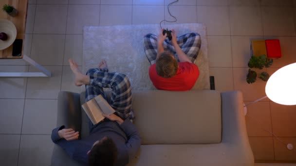 Top shot de cara em roupas vermelhas jogando videogame com joystick e outro lendo um livro na sala de estar . — Vídeo de Stock