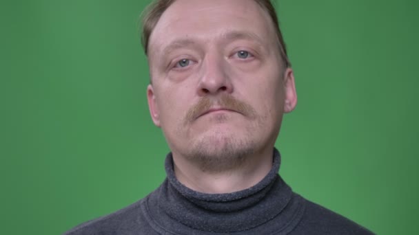 Middle-aged man with beard watching into camera with no emotions inclines head rightwards on green background. — Stock Video