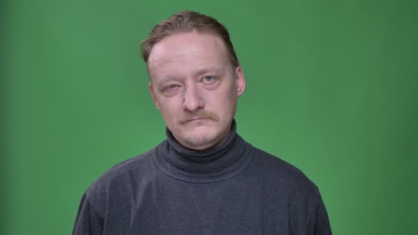 Blonder Mann mittleren Alters in grauem Pullover blickt nachdenklich in die Kamera und zwinkert ihr auf grünem Hintergrund zu. — Stockvideo