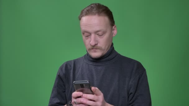 Plavovlasý důchodce v šedém pulovníku, který pozorně pozoruje smartphone a emocionálně reaguje na zelené pozadí. — Stock video