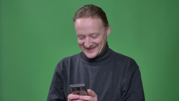 Blonde Rentnerin im grauen Pullover schaut aufmerksam ins Smartphone und lacht vor grünem Hintergrund fröhlich. — Stockvideo