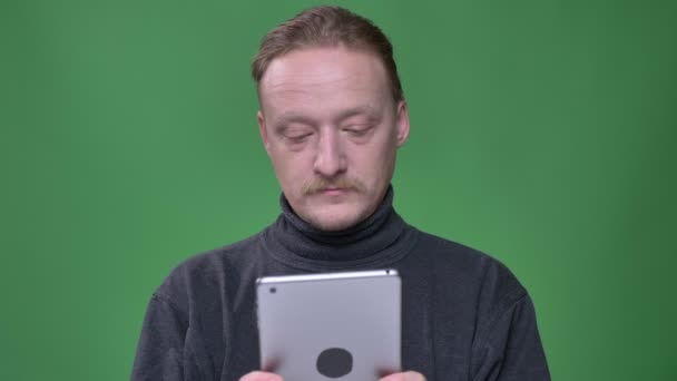 Pensionista rubia en jersey gris mirando atentamente en la tableta y reaccionando emocionalmente sobre fondo verde . — Vídeos de Stock