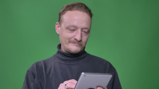 Pensionista rubia en jersey gris trabajando con atención en la tableta y mira en la cámara sobre fondo verde . — Vídeos de Stock